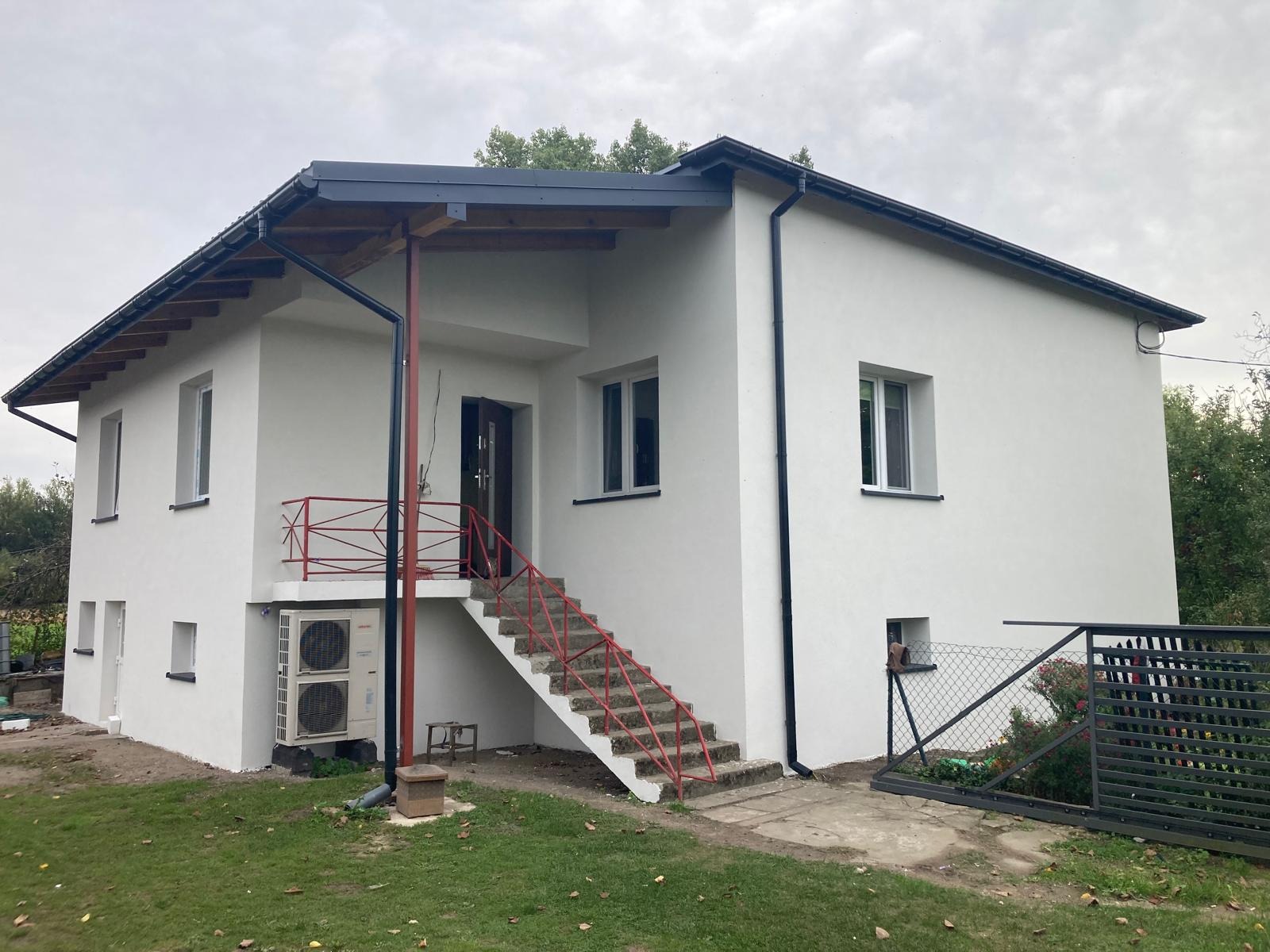 Dwupiętrowy dom po renowacji elewacji, z zewnętrznymi schodami z czerwoną barierką, klimatyzator na zewnątrz i świeżo pomalowaną białą elewacją.