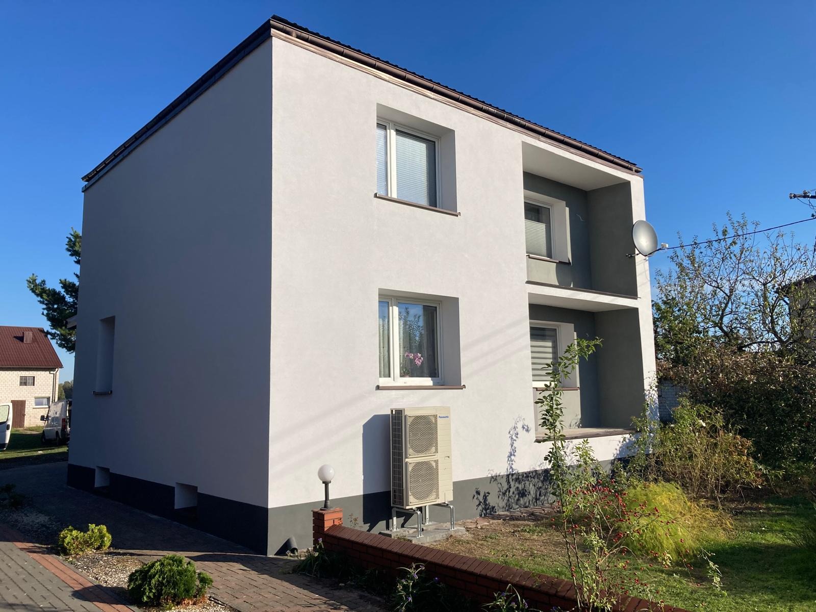 Dwupiętrowy dom z jasnoszarą elewacją, ciemnoszarą podmurówką, balkonami i klimatyzacją na zewnątrz, widziany z poziomu chodnika z kostki.