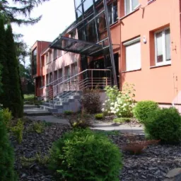 Nowoczesny budynek biurowy z przeszklonym wejściem i zadbanym ogrodem z tujami i korą.