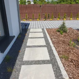 Nowoczesna ścieżka z betonowych płyt prowadząca przez ogród, obłożona czarnym grysem i korą, z widocznym drewnianym ogrodzeniem w tle.