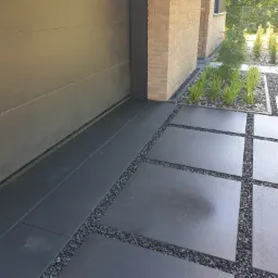 Nowoczesna nawierzchnia z szarych płyt betonowych oddzielonych drobnym żwirem, przylegająca do ściany budynku z jasnej cegły i bramy garażowej, z elementami zieleni w tle.