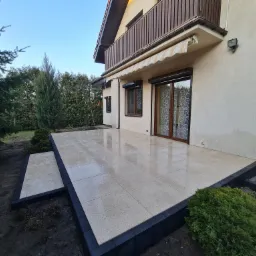 Nowoczesny taras z jasnych płyt z ciemną obwódką, przylegający do domu z balkonem i markizą, otoczony zielenią.