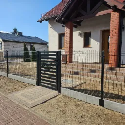 Nowoczesne ogrodzenie panelowe z furtką w kolorze grafitowym, z podmurówką betonową, prowadzące do domu z ceglanymi filarami werandy i kostką brukową, w tle budynek w stanie surowym.