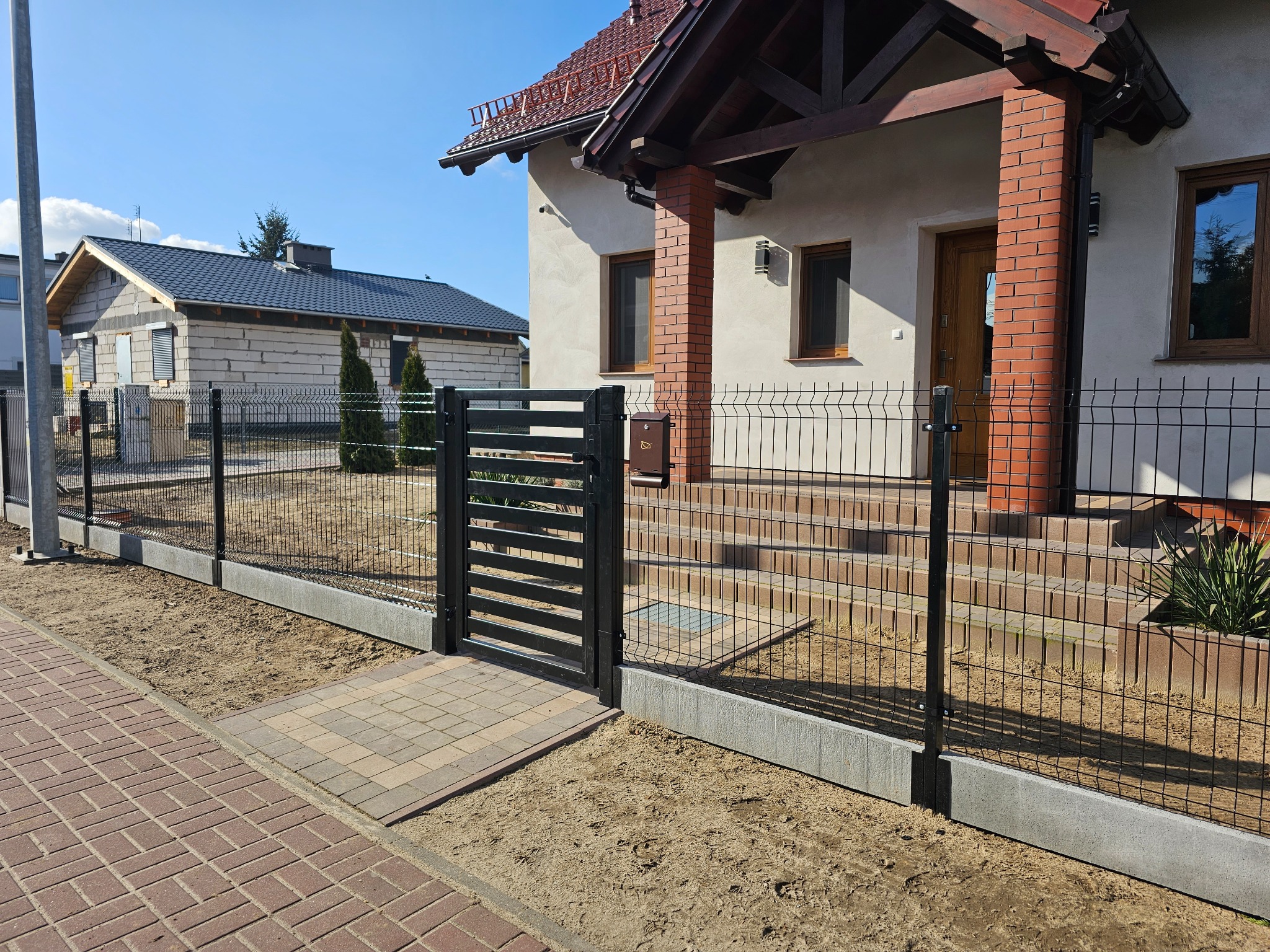 Nowoczesne ogrodzenie panelowe z furtką w kolorze grafitowym, z podmurówką betonową, prowadzące do domu z ceglanymi filarami werandy i kostką brukową, w tle budynek w stanie surowym.