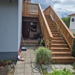 PPUH TEUBNER - Drewniane schody zewnętrzne z balustradą prowadzące do drzwi wejściowych, z narzędziami na stopniach i roślinnością w otoczeniu. Elementy wykończeniowe w kolorze naturalnego drewna.