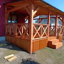 Drewniana, nowa altana z balustradą w kształcie krzyżyków, przylegająca do domu z czerwonym dachem. Widoczne schody wejściowe i dwie deski na ziemi.