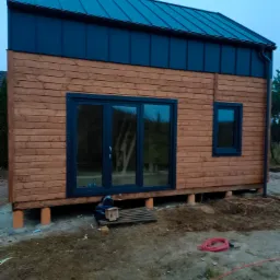 Surowy dom szkieletowy z drewnianą elewacją i ciemnym dachem, podniesiony na betonowych fundamentach. Widoczne okno i drzwi, teren wok&oacute;ł w trakcie prac.