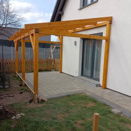 PPUH TEUBNER - Drewniana pergola z jasnego drewna, częściowo zamontowana przy budynku z wyjściem na taras z kostki. Widoczne elementy montażowe i teren wokół w trakcie prac.
