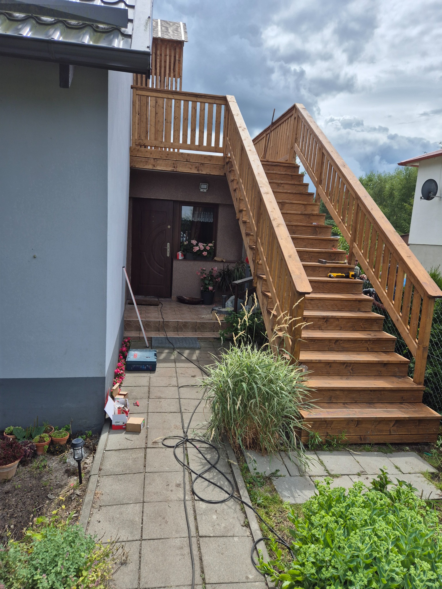 Drewniane schody zewnętrzne z balustradą prowadzące do drzwi wejściowych, z narzędziami na stopniach i roślinnością w otoczeniu. Elementy wykończeniowe w kolorze naturalnego drewna.