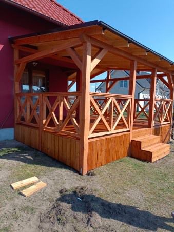 Drewniana, nowa altana z balustradą w kształcie krzyżyków, przylegająca do domu z czerwonym dachem. Widoczne schody wejściowe i dwie deski na ziemi.