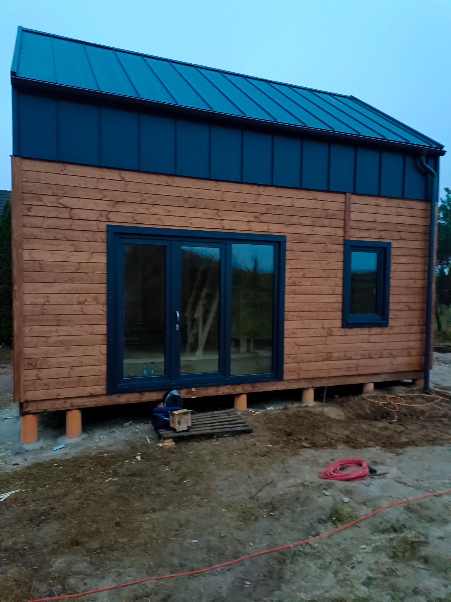 Surowy dom szkieletowy z drewnianą elewacją i ciemnym dachem, podniesiony na betonowych fundamentach. Widoczne okno i drzwi, teren wokół w trakcie prac.