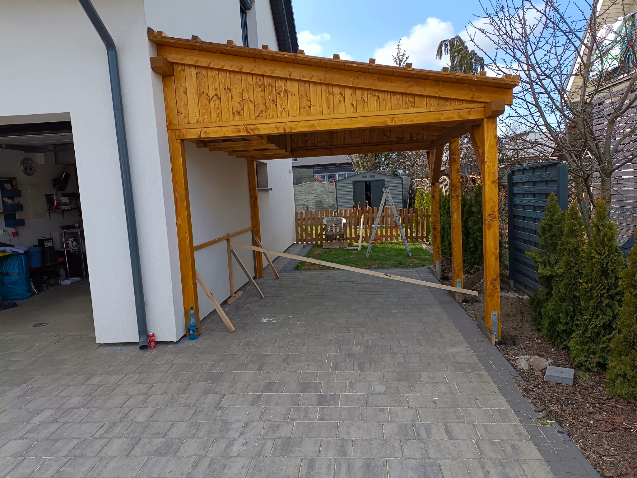 Drewniana wiata garażowa przylegająca do białego budynku, z widoczną konstrukcją dachu i drewnianymi słupami. Na podjeździe kostka brukowa, w tle ogród z zielenią.