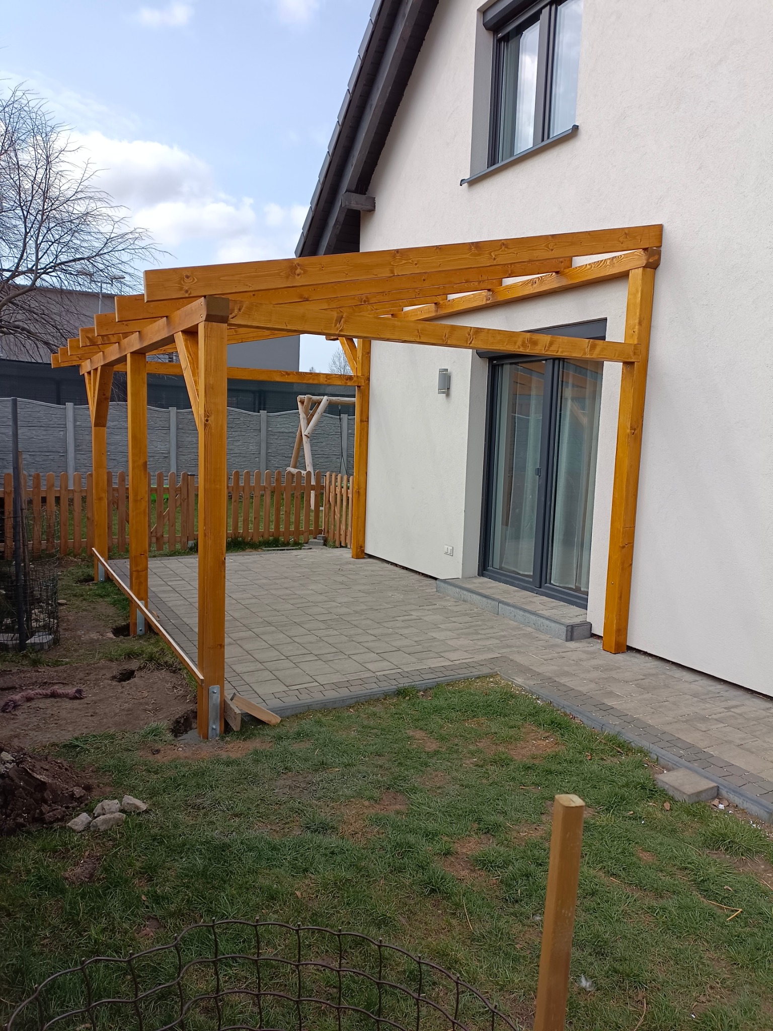 Drewniana pergola z jasnego drewna, częściowo zamontowana przy budynku z wyjściem na taras z kostki. Widoczne elementy montażowe i teren wokół w trakcie prac.