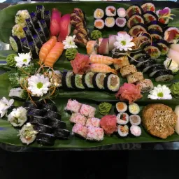 Bogato udekorowany talerz z różnorodnym sushi, ułożonym na liściach bananowca, z dekoracjami z kwiatów i wasabi.