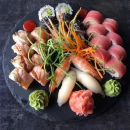 Zestaw sushi z r&oacute;żnymi rodzajami ryb, krewetek i warzyw ułożony na okrągłym, ciemnym talerzu z łupka, udekorowany wasabi, imbirem i nitkami marchewki.