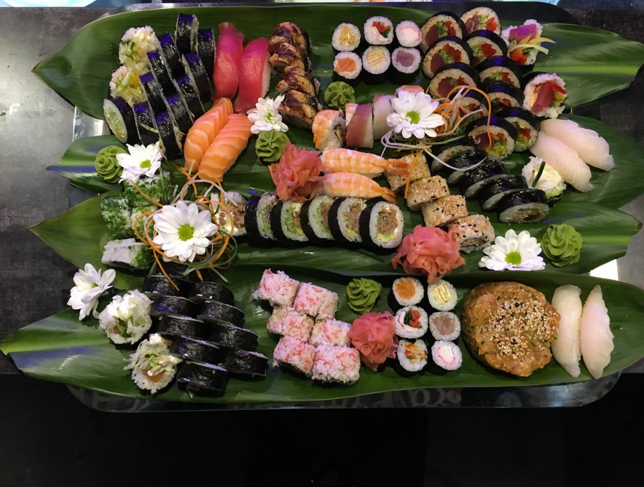 Bogato udekorowany talerz z różnorodnym sushi, ułożonym na liściach bananowca, z dekoracjami z kwiatów i wasabi.