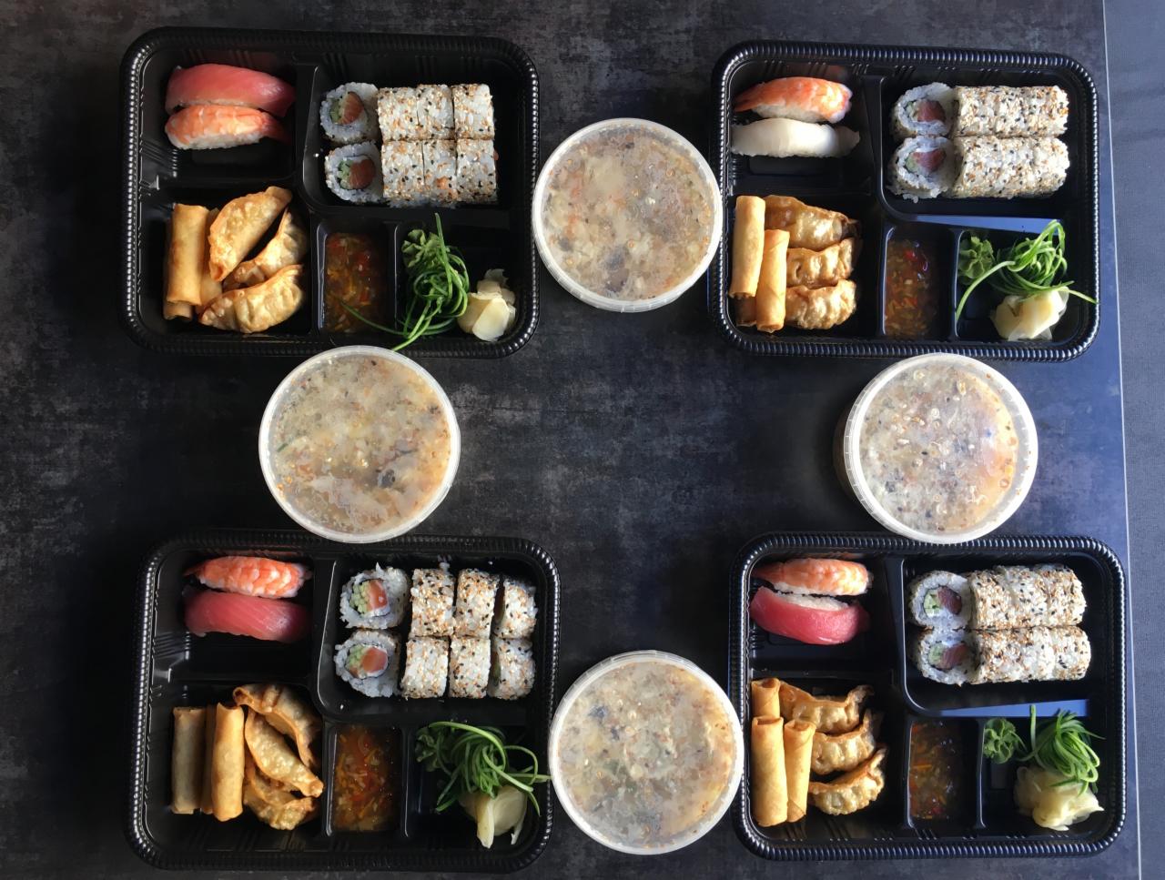 Cztery czarne pudełka cateringowe z sushi, sajgonkami, pierożkami gyoza, sosem i imbirem, uzupełnione zupą w okrągłych pojemnikach, ułożone symetrycznie na ciemnym tle.