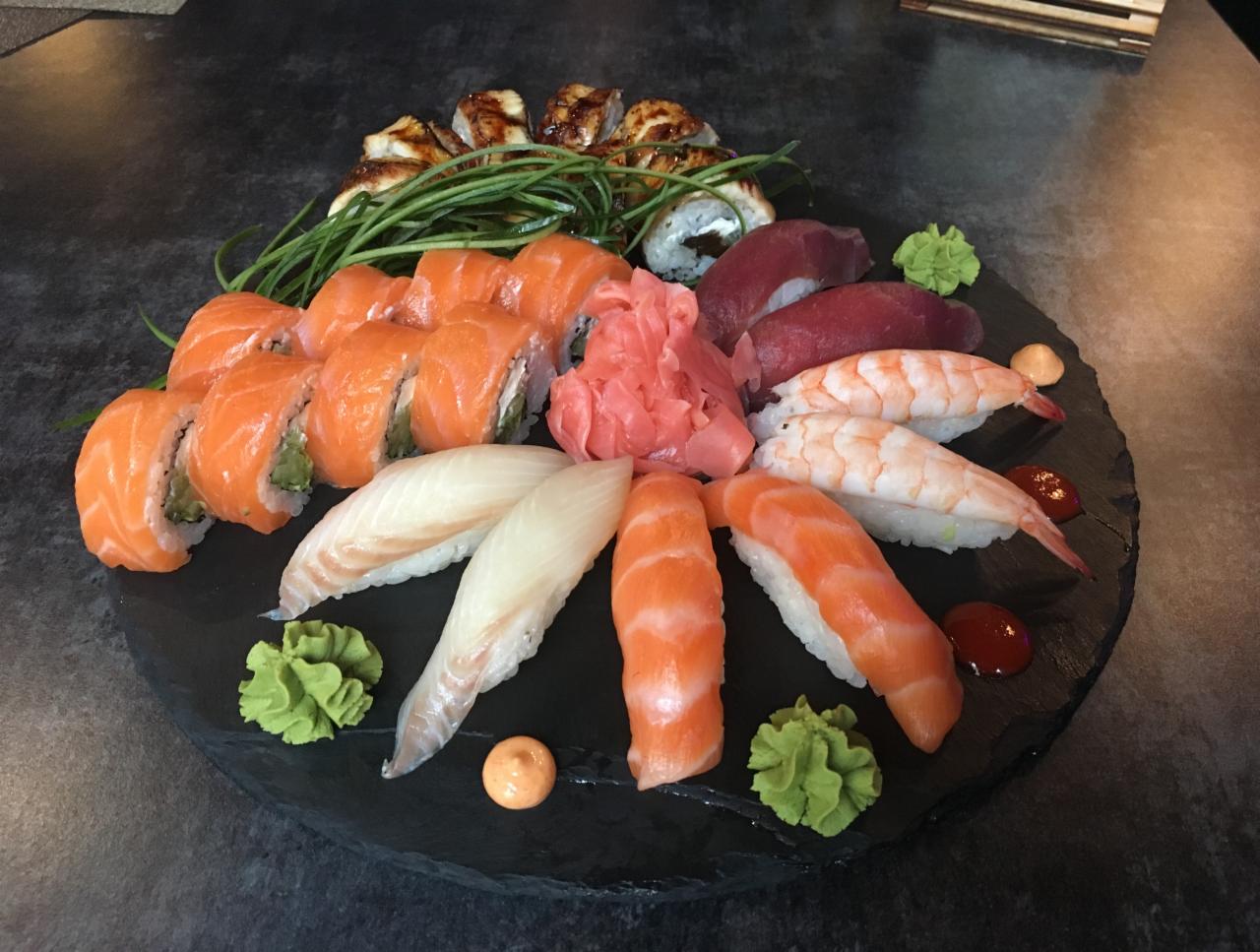 Zestaw sushi z różnymi rodzajami ryb, krewetek i dodatkami, ułożony na czarnym, okrągłym talerzu z łupka, z widocznymi kawałkami imbiru i wasabi.