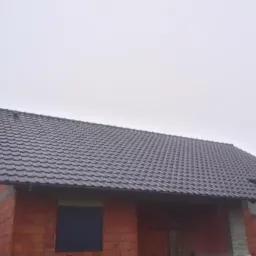 Nowy, ciemny dach z dachówki ceramicznej na budynku w trakcie budowy, widoczne czerwone cegły i szare, ponure niebo.