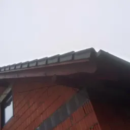 Fragment dachu z czarną dachówką ceramiczną, widoczny z dołu pod kątem, z ceglaną ścianą budynku w tle i pochmurnym niebem.