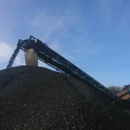 Czarny węgiel kamienny wysypywany z taśmociągu na hałdę, widoczne urządzenia do transportu i składowania węgla, błękitne niebo w tle.