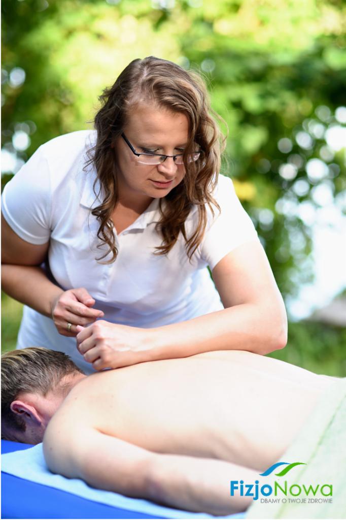 Kobieta w białym uniformie wykonuje masaż pleców pacjenta na zewnątrz, na niebieskim stole przykrytym zielonym ręcznikiem, logo firmy widoczne w prawym dolnym rogu.