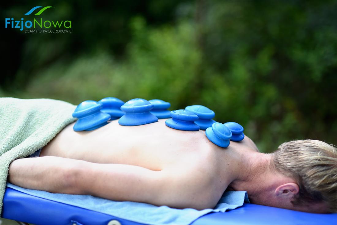 Pacjent leżący na brzuchu z niebieskimi bańkami próżniowymi na plecach, widoczne logo FizjoNowa w górnym rogu, tło z zielenią.