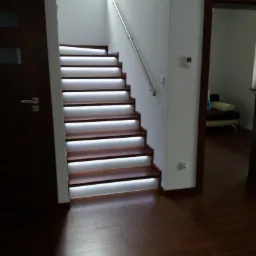 Nowoczesne schody drewniane z podświetleniem LED na każdym stopniu, srebrna poręcz, widok z dołu na brązową podłogę i drzwi.