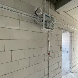 Wewnętrzna instalacja elektryczna w surowym budynku z białych bloczków, widoczne kable i puszki elektryczne, prowadzone pod sufitem i w dół ściany, stan surowy.