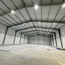 M - INSTAL Michał Lenarczyk - Wnętrze hali magazynowej z widoczną konstrukcją stalową, oświetleniem LED i betonową posadzką. Ujęcie z dołu podkreśla przestronność i wysokość obiektu. Częściowo widoczne materiały budowlane.