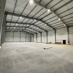 M - INSTAL Michał Lenarczyk - Wnętrze hali magazynowej z oświetleniem LED pod sufitem, widoczne stalowe konstrukcje i betonowa posadzka. Częściowo widoczne materiały budowlane.