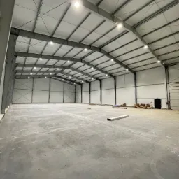 Wnętrze hali magazynowej z oświetleniem LED pod sufitem, widoczne stalowe konstrukcje i betonowa posadzka. Częściowo widoczne materiały budowlane.