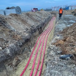 M - INSTAL Michał Lenarczyk - Rów z ułożonymi, różowymi kablami w ziemi. W tle widoczny pracownik w pomarańczowej kamizelce oraz sprzęt budowlany. Kabel zabezpieczony czarnymi opaskami.