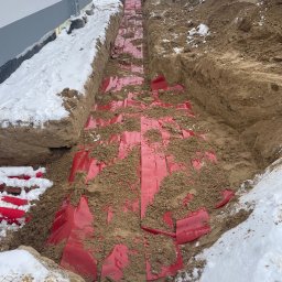 M - INSTAL Michał Lenarczyk - Wykop w ziemi zabezpieczony czerwoną folią, z widocznym pracownikiem w oddali i rurami przy budynku. Zima, śnieg na ziemi.