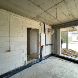 M - INSTAL Michał Lenarczyk - Surowy stan domu z instalacją elektryczną: białe kable i rury prowadzone po ścianach z betonu komórkowego, skrzynka rozdzielcza, widok na zewnątrz budynku.