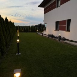 M - INSTAL Michał Lenarczyk - Oświetlenie ścieżki wzdłuż trawnika i żywopłotu, z nowoczesnymi lampami ogrodowymi, tworzące nastrojowy klimat przy domu z białymi ścianami i elementami w kolorze bordo.