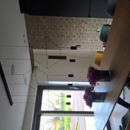 Nowoczesna jadalnia z minimalistyczną lampą wiszącą z czarnymi elementami, drewnianym stołem i kolorowymi krzesłami. Lampa z cienkimi przewodami i podłużnymi oprawami, zawieszona nad stołem z dwoma...