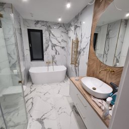 Tomasz Pieczyński - Nowoczesna łazienka z marmurowymi płytkami, owalną umywalką na drewnianym blacie, złotą armaturą i grzejnikiem drabinkowym, kabina prysznicowa ze szklanymi drzwiami.