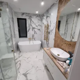 Nowoczesna łazienka z marmurowymi płytkami, owalną umywalką na drewnianym blacie, złotą armaturą i grzejnikiem drabinkowym, kabina prysznicowa ze szklanymi drzwiami.