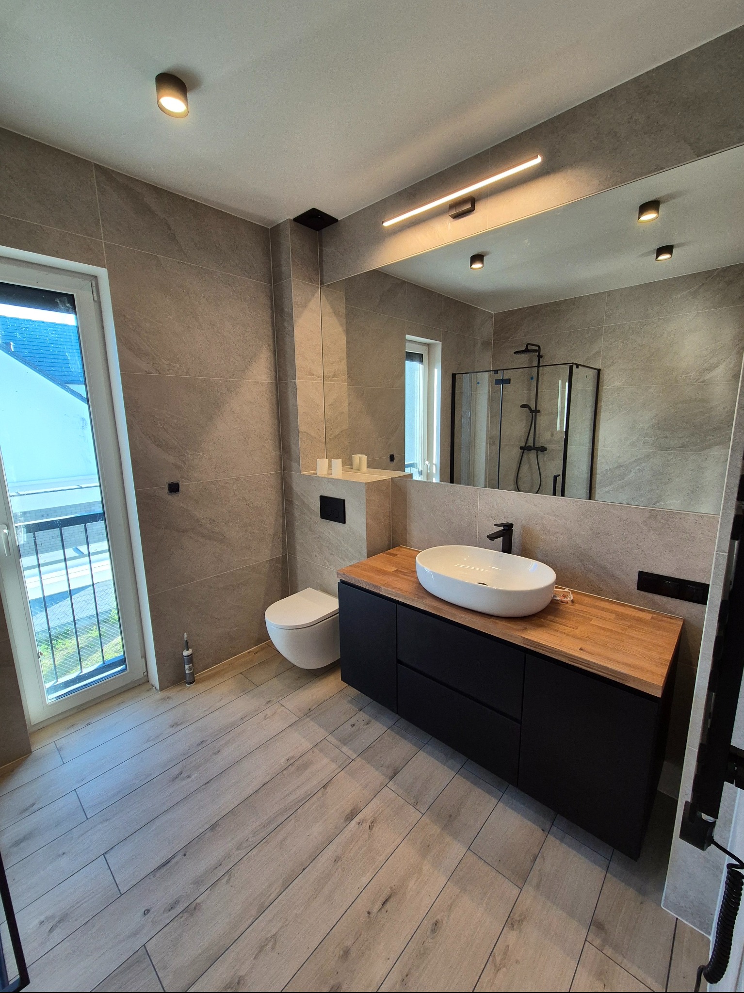 Nowoczesna łazienka z płytkami imitującymi kamień, drewnopodobną podłogą, owalną umywalką na blacie z jasnego drewna i kabiną prysznicową za lustrem.