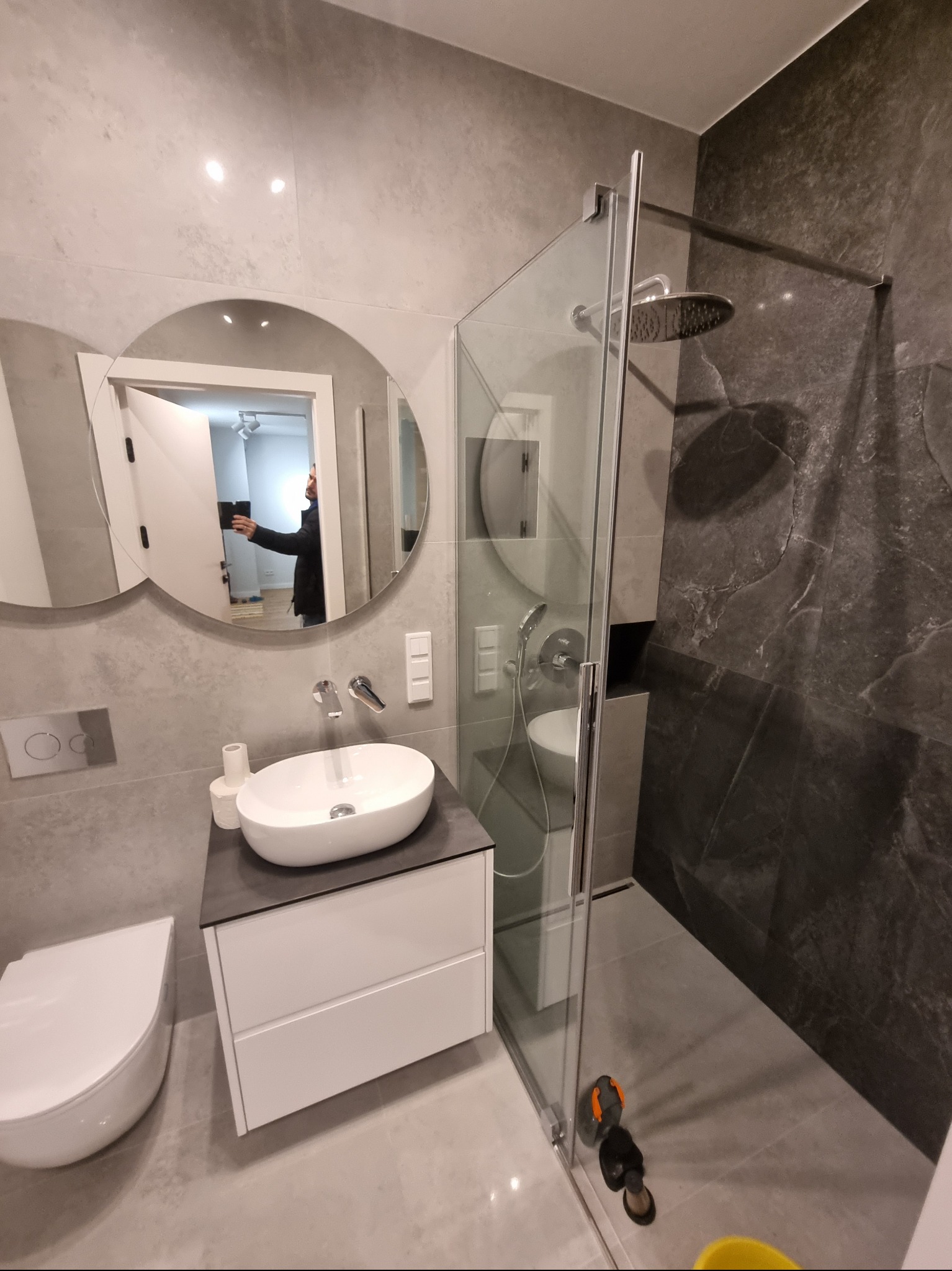 Nowoczesna łazienka z owalnym lustrem, białą umywalką nablatową, kabiną prysznicową typu walk-in z deszczownicą i szarą okładziną ścienną imitującą marmur, widok z perspektywy osoby robiącej...