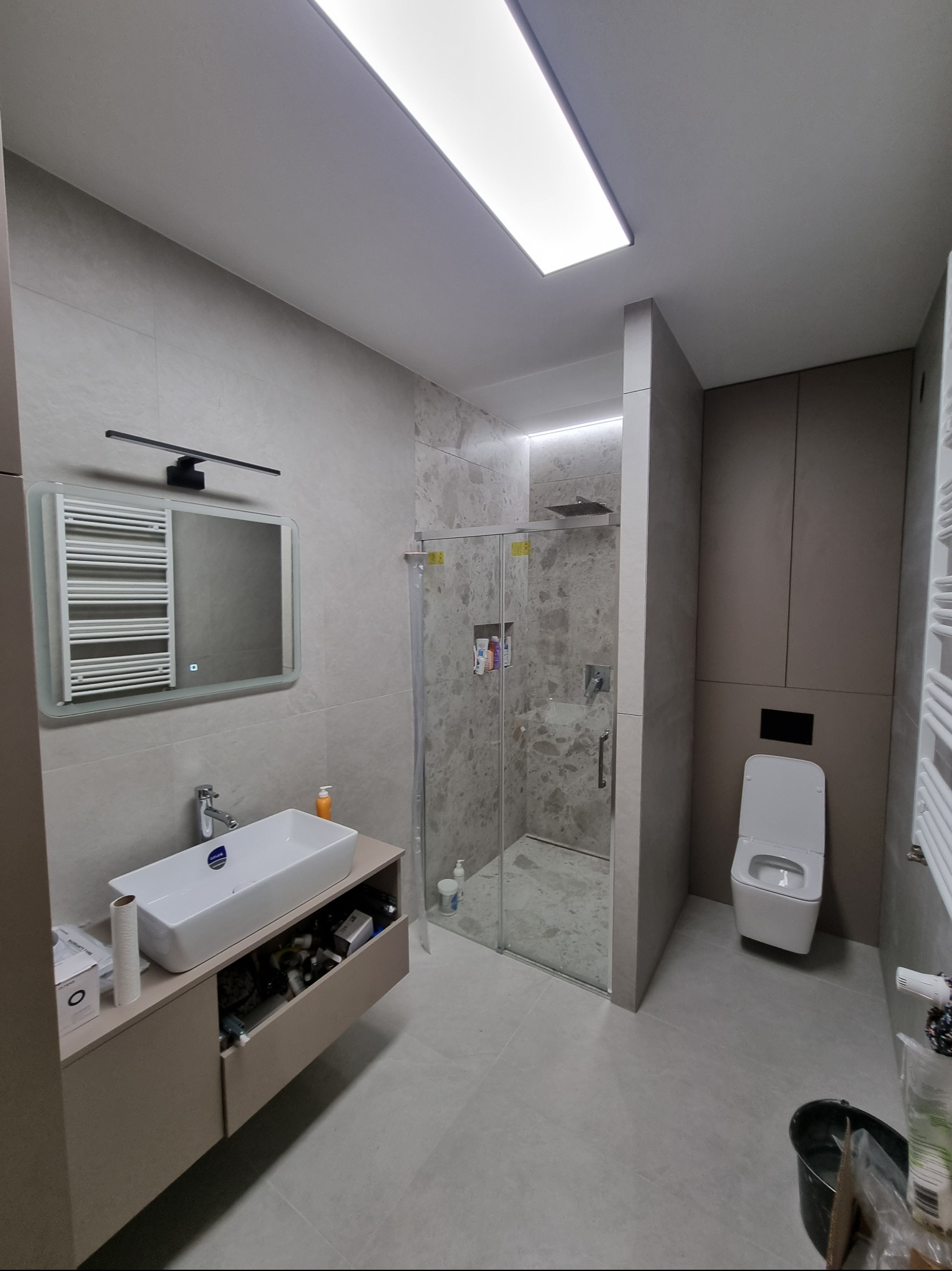 Nowoczesna łazienka z płytkami imitującymi marmur, kabiną prysznicową typu walk-in, podwieszaną toaletą i umywalką nablatową z szafką w odcieniach beżu, oświetlona ledowym panelem sufitowym.