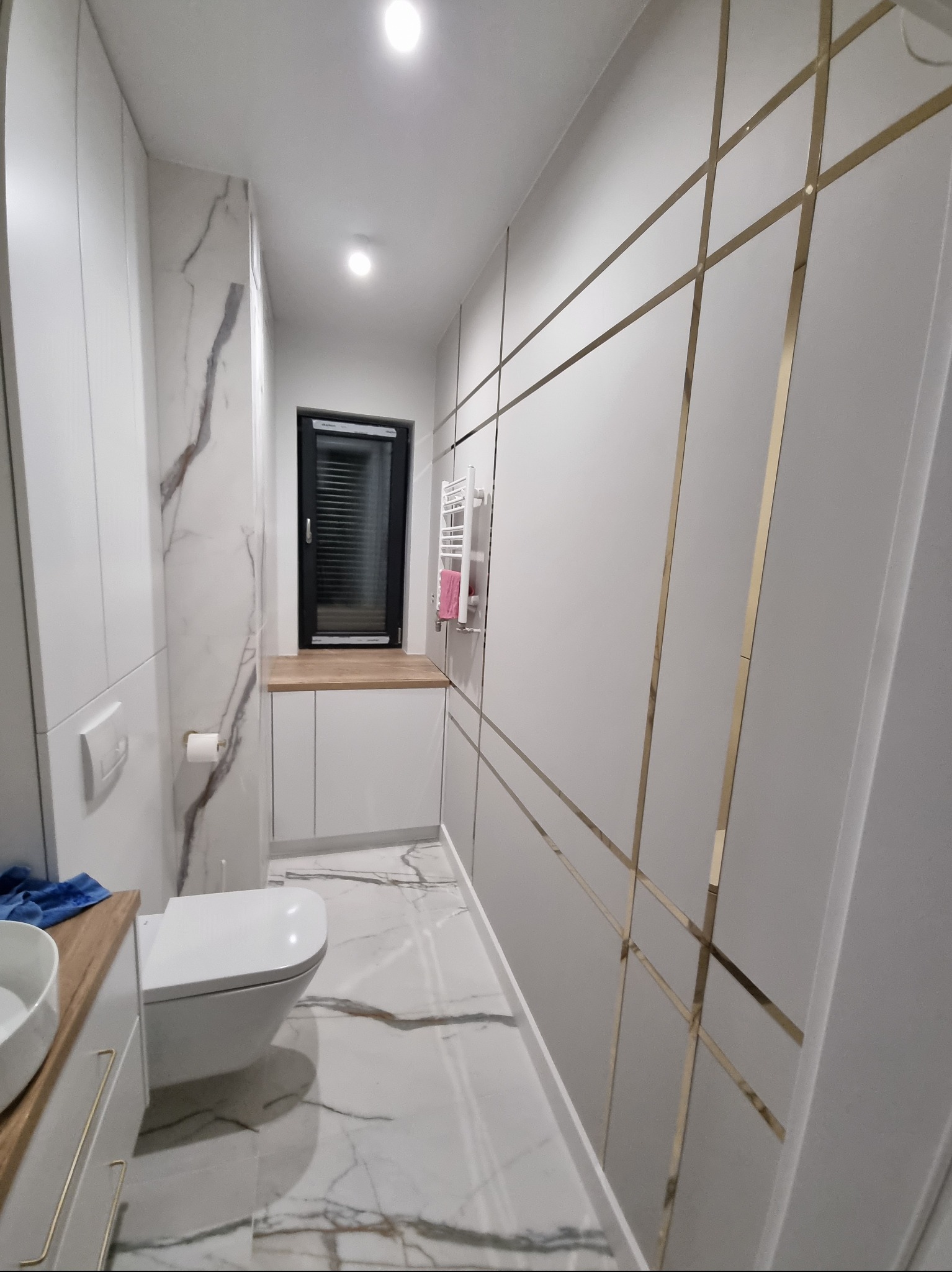 Nowoczesna łazienka z białymi płytkami imitującymi marmur na podłodze i ścianie, biała toaleta podwieszana, szafka z umywalką z drewnianym blatem, ściana zdobiona złotymi listwami tworzącymi...