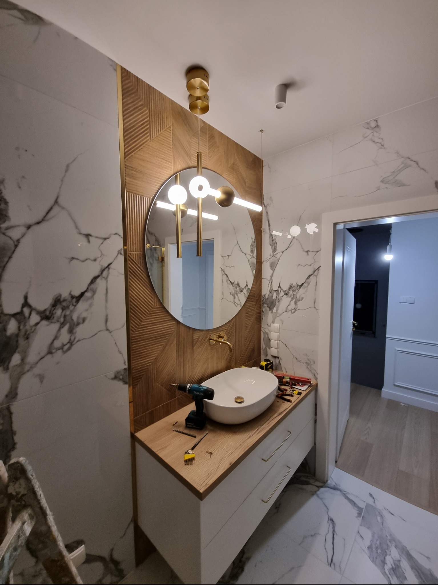 Montaż umywalki nablatowej w łazience z marmurowymi płytkami i dekoracyjnym oświetleniem, widoczna wiertarka i narzędzia na blacie szafki.