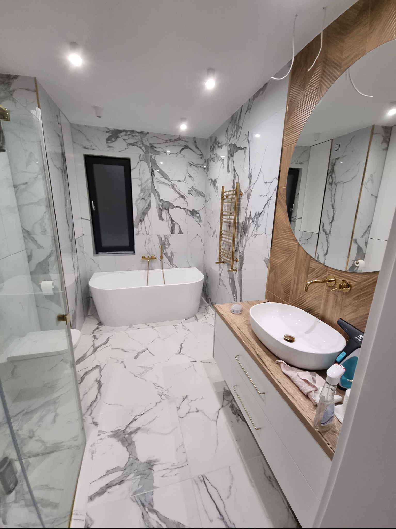 Nowoczesna łazienka z marmurowymi płytkami, owalną umywalką na drewnianym blacie, złotą armaturą i grzejnikiem drabinkowym, kabina prysznicowa ze szklanymi drzwiami.