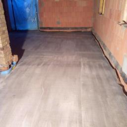 Ubik Bud - Surowy betonowy podkład podłogowy w nowo budowanym domu z czerwonej cegły, widoczne zabezpieczenie folią drzwi wejściowych.