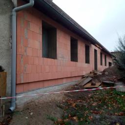 Ubik Bud - Ściana budynku w trakcie budowy z pustaków ceramicznych, widoczne otwory okienne, rynna spustowa, teren budowy zabezpieczony taśmą.