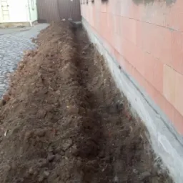 Wykopany wąski rów wzdłuż ściany budynku z czerwonej cegły, widoczny fragment fundamentu, na górze kostka brukowa i ogrodzenie.