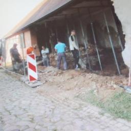 Ubik Bud - Ekipa budowlana zabezpiecza uszkodzoną ścianę budynku za pomocą metalowych podpór; w tle widoczny gruz i narzędzia, a na pierwszym planie kostka brukowa i bariera ostrzegawcza.