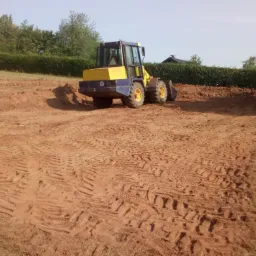 Żółto-niebieska ładowarka kołowa na terenie przygotowywanym pod budowę, widoczne ślady opon w ziemi.
